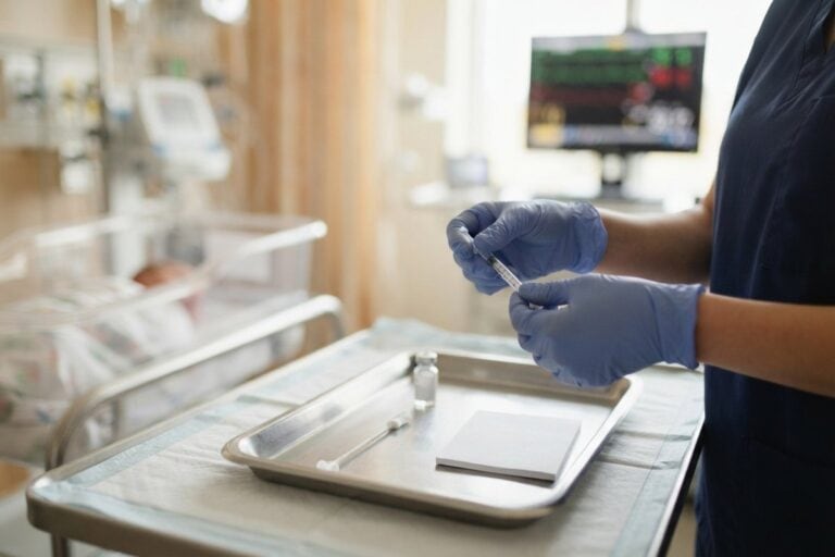 Mains gantées préparant une seringue au-dessus d'un plateau métallique, près d'un berceau de nouveau-né en maternité