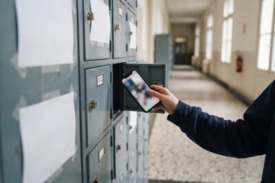 Main d'un adolescent déposant un smartphone dans un casier métallique ouvert, dans un couloir de lycée